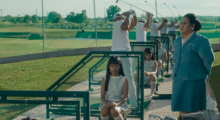 A line of golfers hoist their clubs mid-swing on a driving range. A young woman in uniform sits nearby, ready to put golf balls on tees for the golfers. Her manager, an older woman in a blazer, looks on at the scene.
