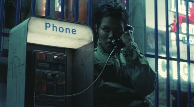 Teyana Taylor has short black hair and wears a khaki jacket. She holds a payphone to her ear and looks off in the distance.
