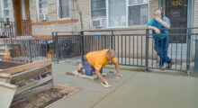 A burly man in a bright yellow shirt and cutoff jean shorts smooths wet concrete on a sidewalk while a balding man smokes a cigarette and looks on.