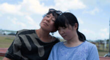 Set against a blue sky, two sisters, both Asian adoptees, lean their heads against each other.