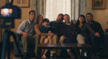 An extended family sits on a couch for a family photo; they make funny faces for the camera.