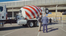 Two men dressed in business attire stand in front of a large concrete mixer, which is decorated by a red candy cane stripe.