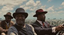 Three Black men wearing 1920s period garb sit in a hoodless car and drive amid a blue, cloud-flecked sky.