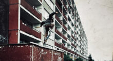 A row of brutalist apartment buildings stand behind a young man as he does a kickflip off of a railing on a skateboard.