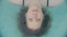 A woman lies on her back in clear blue water, her red hair forms a halo around her head. Her bright blue eyes look straight above her.