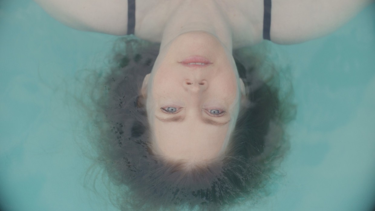 A woman lies on her back in clear blue water, her red hair forms a halo around her head. Her bright blue eyes look straight above her.