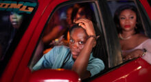 A Nigerian woman in a blue blouse puts her hand atop her cropped hair and drives a red taxi. A few women are seated in the back.