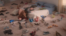 A woman wearing a gray mesh long sleeve, a plaid knee-length skirt, sheer gray knee-high socks and mismatched shoes lays her head on her bed while the rest of her body lies on the floor. She is surrounded by shoes, which are scattered all over the pink carpet.
