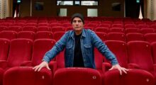 An older Iranian man stands in the middle in a row of red seat at a movie theater. He wears a black t-shirt, blue denim jacket and black beanie.