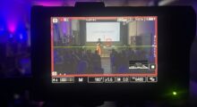 A camera monitor records a woman on stage, wearing mustard pants and a black shirt, giving a presentation. The screen behind her reads "I Said What I Said."