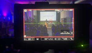 A camera monitor records a woman on stage, wearing mustard pants and a black shirt, giving a presentation. The screen behind her reads "I Said What I Said."