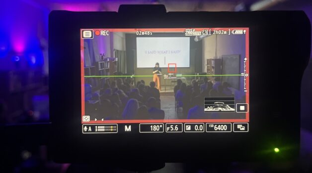 A camera monitor records a woman on stage, wearing mustard pants and a black shirt, giving a presentation. The screen behind her reads "I Said What I Said."