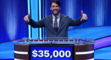 A man with brown hair and glasses wears a gray suit and purple, blue and white floral tie. He stands at a Jeopardy! podium and points down to the screen which shows $35,000 of winnings.