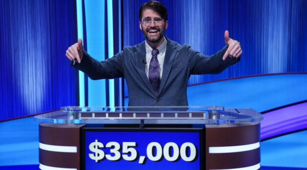 A man with brown hair and glasses wears a gray suit and purple, blue and white floral tie. He stands at a Jeopardy! podium and points down to the screen which shows $35,000 of winnings.