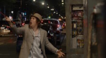 A man wearing a beige blazer, fedora and button-up clutches an orange extension cord and raises it in the air. He makes a shocked facial expression.