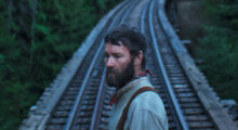 A rugged man wearing dingy early 19th century garb stands in front of wooden train tracks.