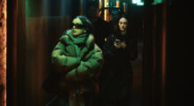 Pop star Charli XCX wears thick black sunglasses and a puffer jacket as she walks with her arms crossed down a dark, green-lighted hallway. A young woman follows closely behind with a tablet in hand.