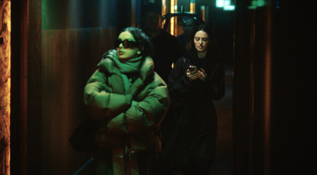 Pop star Charli XCX wears thick black sunglasses and a puffer jacket as she walks with her arms crossed down a dark, green-lighted hallway. A young woman follows closely behind with a tablet in hand.