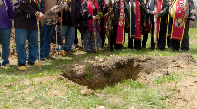 A group of men with shovels surround an open grave in a grassy field. None of the men's faces are shown and some wear ceremonial red sashes.