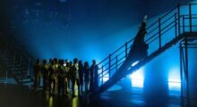 A pop star wearing an ornate headpiece and donning a long cape ascends a flight of stairs backstage; behind her are back-up dancers. They are all washed in blue and yellow stage lights, cast in shadow.