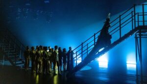 A pop star wearing an ornate headpiece and donning a long cape ascends a flight of stairs backstage; behind her are back-up dancers. They are all washed in blue and yellow stage lights, cast in shadow.
