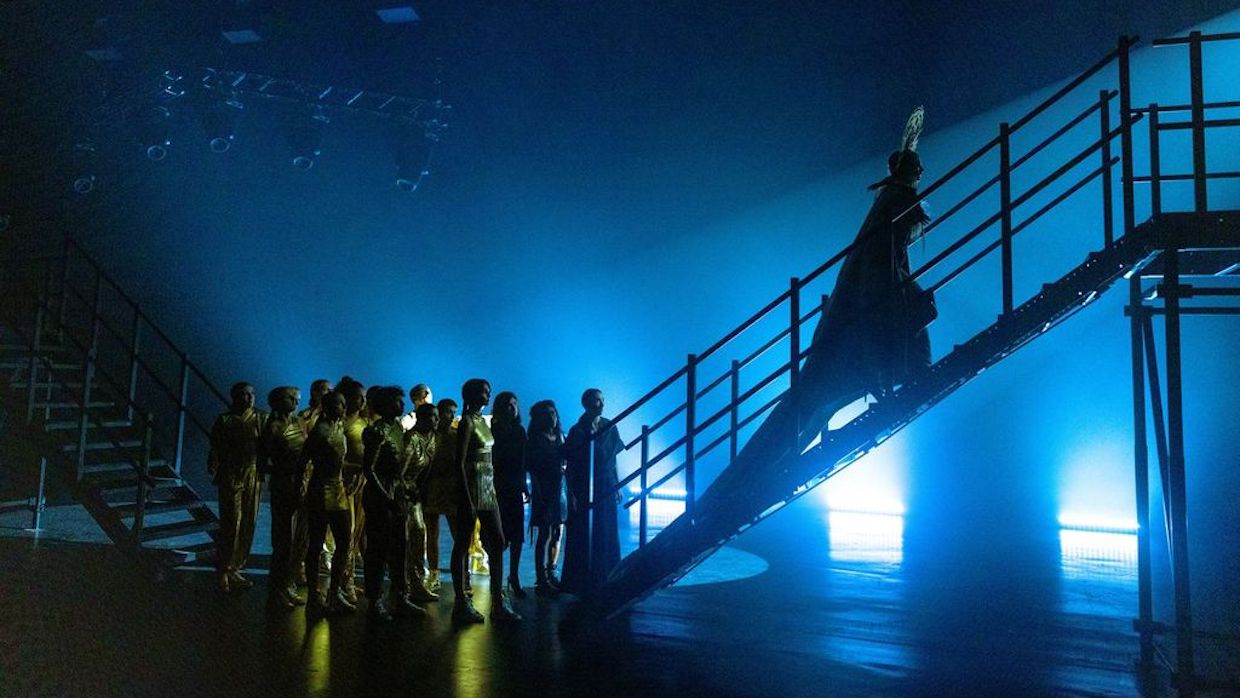 A pop star wearing an ornate headpiece and donning a long cape ascends a flight of stairs backstage; behind her are back-up dancers. They are all washed in blue and yellow stage lights, cast in shadow.