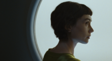 A white woman with a brown pixie cut stands in front of a cloudy, gray window. Her head is turned so you see only her right profile.