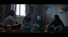 Two adult siblings and a 13-year-old girl sit in a bedroom. The adult man and the teen sit on a bed with red sheets and a blue comforter. The woman sits on a chair beside the bed. They appear deep in conversation.