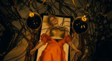 A woman lays on a 19th century surgery table. Her orange silk dress is unbuttoned and electrical wires run through her torso.