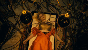 A woman lays on a 19th century surgery table. Her orange silk dress is unbuttoned and electrical wires run through her torso.
