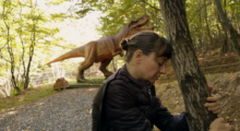 A woman crouches in a wooded area; a plastic t-rex looms behind her.