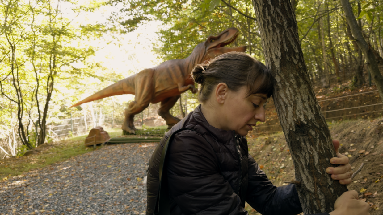 A woman crouches in a wooded area; a plastic t-rex looms behind her.