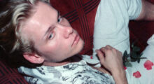 A 25-year-old man with peach fuzz and slicked back blond hair sits askew on a couch. He wears sun-bleached denim and a white shirt with a black floral design. Rose petals sit in his lap.