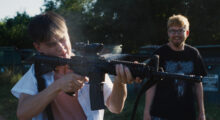 Two young white men stand in front of a dumpster in a field. One wears a white t-shirt and aims a rifle in the distance. The other stands to the side and smiles.