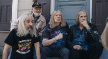Rob Reiner sits behind the cast of This Is Spinal Tap. They are all older white men with graying hair. Three men sit in fron of Reiner, all wearing band tees and denim. Reiner, sitting behind them on a stoop, wears a khaki jacket and black cap.