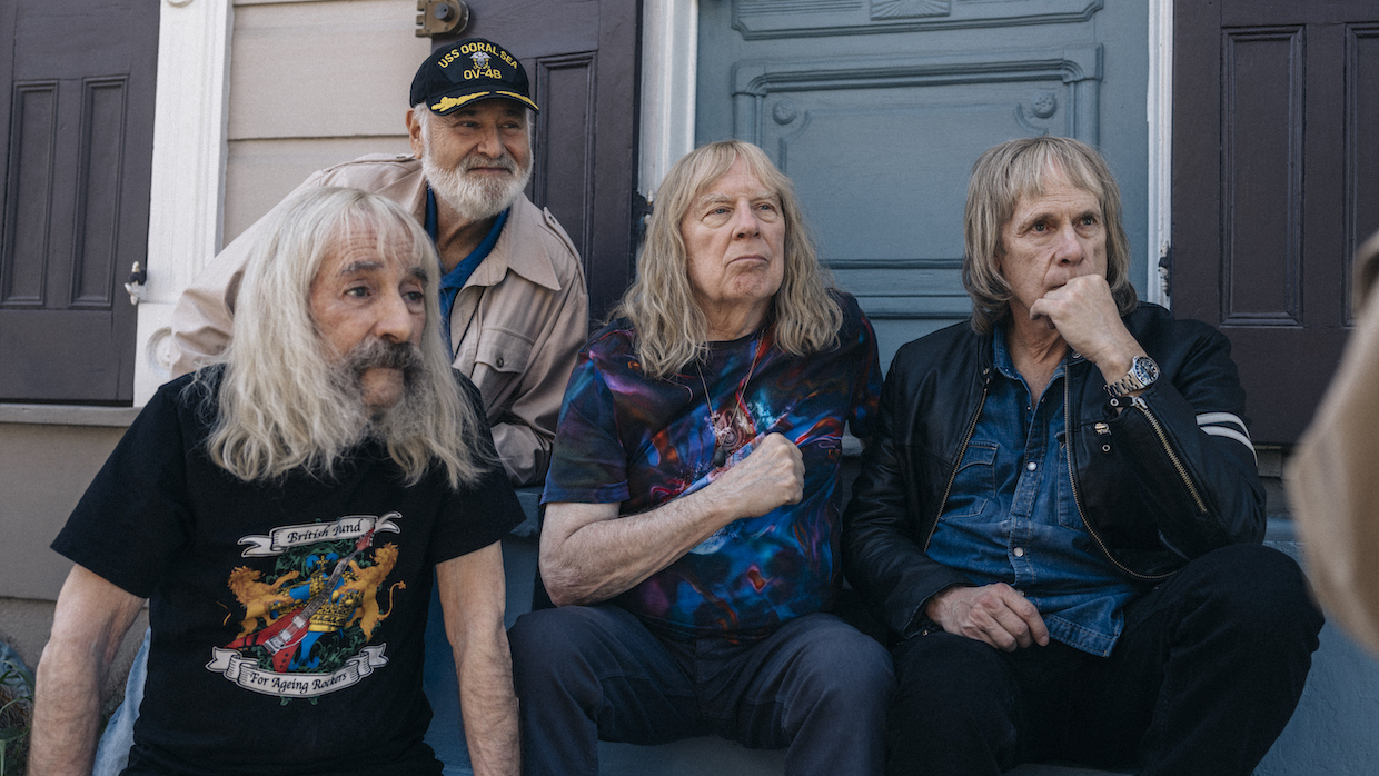 Rob Reiner sits behind the cast of This Is Spinal Tap. They are all older white men with graying hair. Three men sit in fron of Reiner, all wearing band tees and denim. Reiner, sitting behind them on a stoop, wears a khaki jacket and black cap.