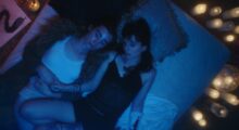 A queer couple lay on a bed surrounded by candles. The the left a pencil drawing of a snake sits by the bed.