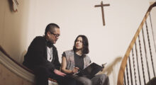A man with headphones sits next to a woman on a winding staircase. She holds a script in her hand. A crucifix is hung above them.