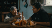 An elderly Indigenous Mexican woman holds a brown-capped mushroom. Her adult granddaughter sits next to her at the kitchen table and listens attentively.