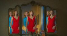 A woman and her body double, both with red hair and one wearing a red and one wearing a blue dress, stare at a mirror.