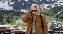 A woman with long, straight blond hair tucks her hair behind her right ear as she walks. She wears black sunglasses, a tan jacket, gray t-shirt and blue jeans. Behind her is a snow-dusted mountain landscape.