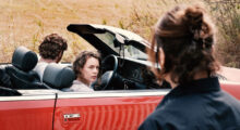 A man drives a red convertible but we only see the back of his head covered in brown hair. A woman with dirty blond curly hair sits in the passenger seat and looks to her right. Another woman stands near the road, we can also only see the back of her head covered in brown hair.