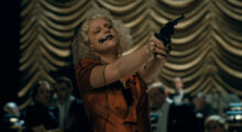A blonde woman with a black ink splotch on her right cheek holds a gun and wears a silk 1930s-style dress