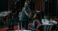 A white man stands in front of a film monitor in a banquet hall. A young Black actress, covered in fake blood, sits in a dining chair and looks up at the footage.
