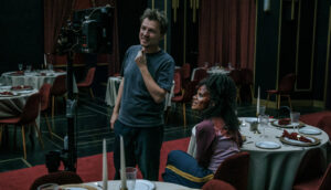 A white man stands in front of a film monitor in a banquet hall. A young Black actress, covered in fake blood, sits in a dining chair and looks up at the footage.