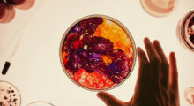 A petri dish contains a swirling mixture of red, purple and orange liquids. Someone's forefinger and thumb touch the dish, an eye dropper and a few jars sit nearby.