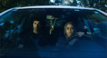 A young Iraqi man and woman sit in a car; through the windshield, we see that she wears a hijab and drives while he wears a black shirt and sits in the passenger seat.