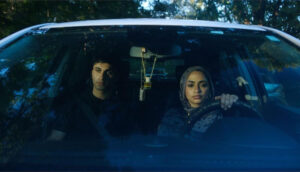 A young Iraqi man and woman sit in a car; through the windshield, we see that she wears a hijab and drives while he wears a black shirt and sits in the passenger seat.