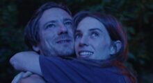 A young man and woman embrace, their faces washed in moonlight as they smile and stare above.