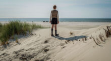 A boy stands on a sandy shore in the summer. He wears 1940s-era clothes, including shorts, a button-up, sweater vest and knee-high socks. His back faces us as he looks out at the blue water on a sunny day.